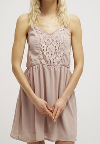 Robe beige sans manches avec des détails en dentelle sur le corsage, une jupe plissée et un col en V. Tissu doux avec une silhouette fluide.