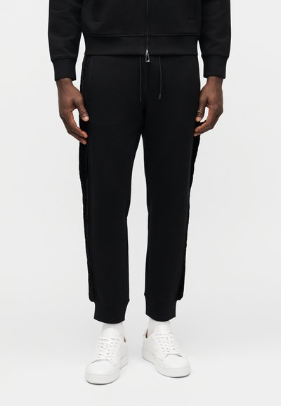 Μαύρο παντελόνι φόρμας από βαμβακερό μείγμα, με υφασμένη πλαϊνή λεπτομέρεια και στενές γραμμές στα πόδια, συνδυασμένο με λευκά αθλητικά παπούτσια και λευκές κάλτσες.