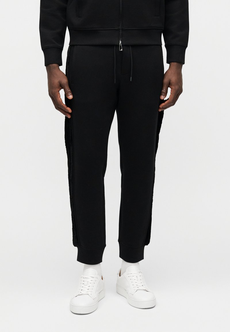 Svarte joggebukser laget av bomullsblanding, med en teksturert sidepanel og innsnevrede ben, kombinert med hvite joggesko og hvite sokker.