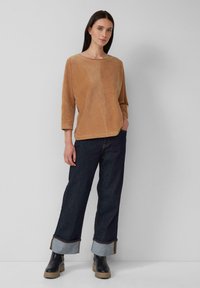 Vrouw in een losse tan kleurige corduroy top, met donkere wide-leg broek met gulden en zwarte chunky-soled enkellaarsjes, staand tegen een grijze muur.