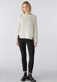 Weißer gerippter Pullover mit Rundhalsausschnitt, der vertikale Nahtdetails und seitliche Reißverschlüsse aufweist, kombiniert mit schwarzen Leggings und glänzenden Schuhen.