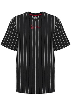 Homme portant un t-shirt noir à rayures verticales, un jean bleu clair ample et des baskets blanches, debout devant un fond gris uni.
