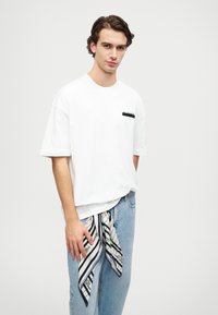 Balts kokvilnas T-krekls ar kabatas dizainu un īsām piedurknēm, kopā ar gaiši ziliem džinsiem. Melnbalts svītrains šalle ir sasieta apvidū.