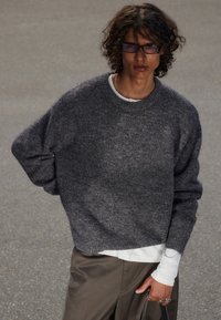 Suéter gris de lana con un corte holgado, escote redondo y hombros caídos, superpuesto a una camisa blanca de manga larga, combinado con pantalones marrones.