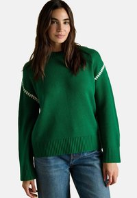 Grüner gestrickter Pullover mit geripptem Saum und Bündchen, mit weißen Akzenten auf den Schultern. Kombiniert mit blauen Jeans.