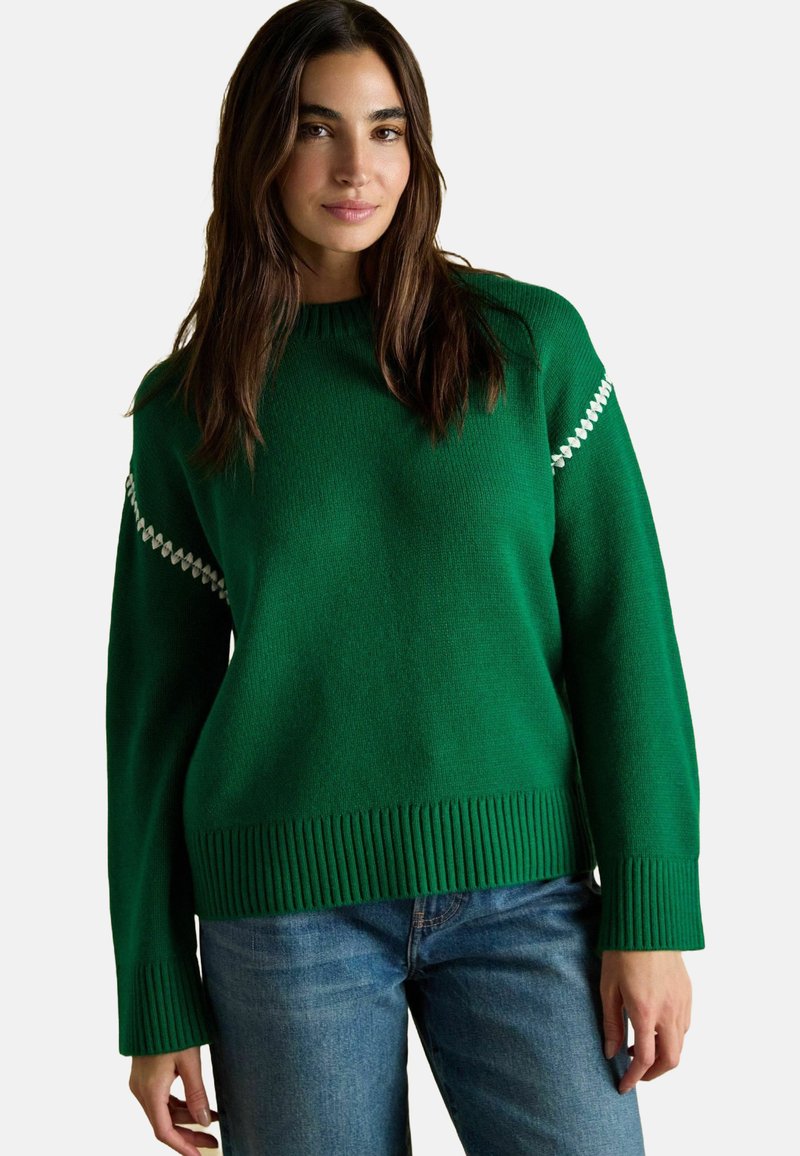 Grüner gestrickter Pullover mit geripptem Saum und Bündchen, mit weißen Akzenten auf den Schultern. Kombiniert mit blauen Jeans.