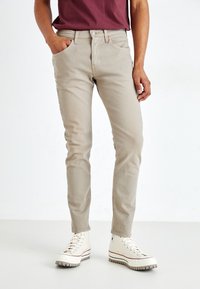 Vaqueros beige claros de corte slim, hechos de una tela texturizada. Bolsillo delantero visible, combinados con zapatillas altas color crema. Exhibición de un atuendo casual.