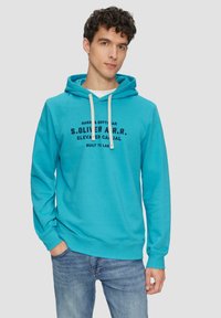 Türkiser Kapuzenpullover mit Kordelzügen, Fronttasche und gedrucktem Textdesign. Kombiniert mit Jeans. Der Stoff wirkt weich und komfortabel.