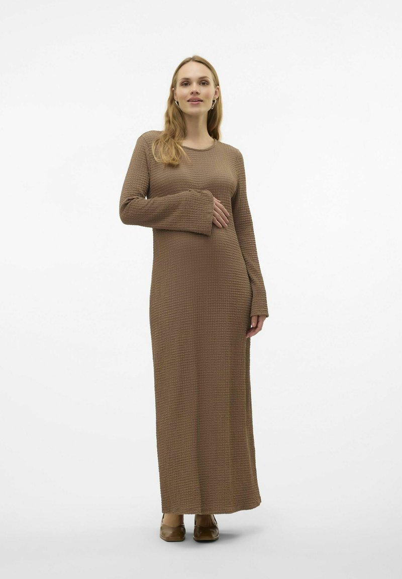 Robe en tricot longue, marron, avec un design texturé ressemblant à des gaufres. Elle possède de longues manches et une coupe décontractée, arrivant sous les chevilles, associée à des chaussures plates.