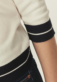 Pull blanc avec poignets côtelés rayés bleu marine et blanc ; texture en maille douce ; coupe ajustée ; porté avec un denim foncé.