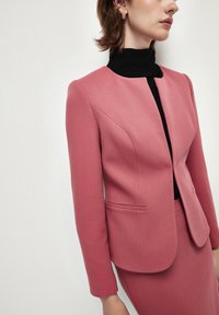 Blazer rosa su misura con una texture liscia, collo rotondo e chiusura frontale nascosta; abbinato a un top nero a collo alto.