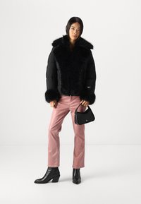 Veste noire rembourrée avec col et poignets en fausse fourrure, assortie à un pantalon en velours côtelé rose et des bottines à talons noires. Tenant un sac à main noir.