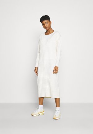 Lang, ribbestrikket kjole i off-white med V-hals og lange ermer. Kombinert med hvite joggesko og ankelstrømper. Enkel, strømlinjeformet design.