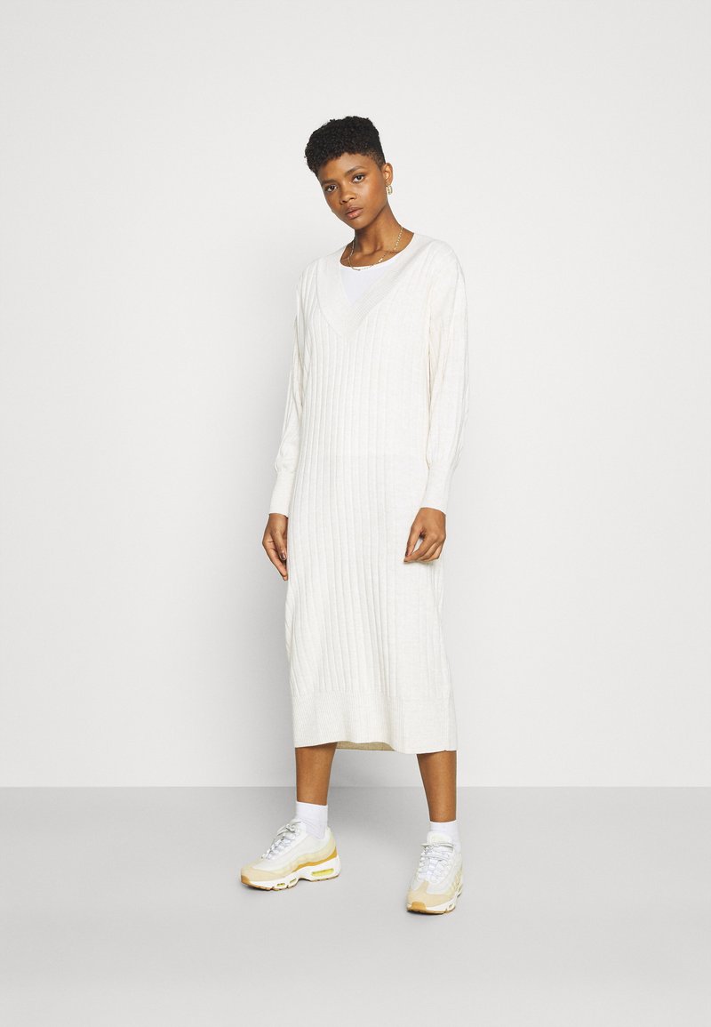 Rochie lungă din tricot cu nervuri, de culoare albă-crem, cu decolteu în V și mâneci lungi. Este asortată cu adidași albaștri și șosete până la gleznă. Design simplu și elegant.