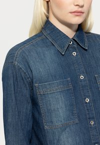 Denim shirt in dark blue with a button-up front, classic collar, and two chest pockets. Features contrasting stitching and glossy buttons.