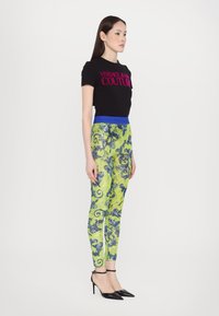 Camiseta negra de manga corta con "Versace Jeans Couture" en rosa. Leggings verde brillante con patrón de paisley azul y cinturilla azul. Tacones negros.