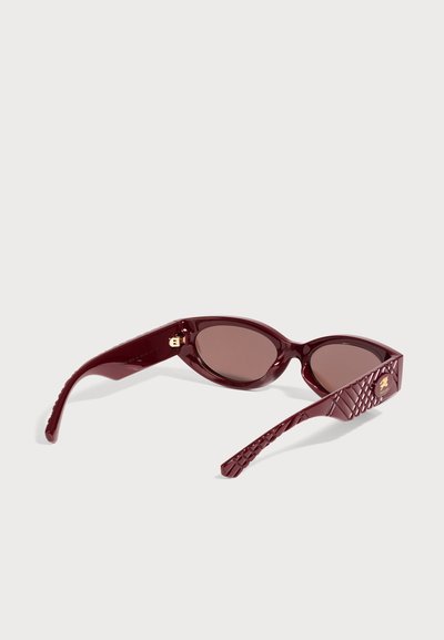 Lunettes de soleil ovales bordeaux, avec des verres teintés, des branches texturées et des détails dorés aux charnières et sur les côtés, sur fond uni.