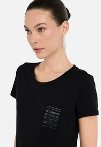 Kvinde med mørkt hår i en knold iført hængende rhinestone øreringe og en sort T-shirt med et lille rhinestone-logo på brystet.
