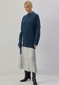 Pull en maille bleu sarcelle foncé avec motifs torsadés, noué à la taille, assorti à une jupe plissée fluide gris clair et des bottes noires montantes jusqu'aux genoux.