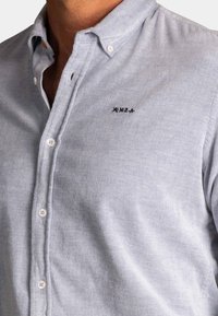 Lichtblauw overhemd met knopen van een gestructureerde stof. Opvallende kenmerken zijn de witte knopen en een klein geborduurd logo op de borst.