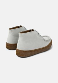 Zapatos casuales en gris claro con un talón alto y punta redonda, que presentan un diseño con cordones y suela marrón texturizada, vistos desde un ángulo posterior.