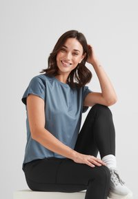Smiling woman with short brown hair in blue athletic shirt and black leggings seated, resting hand on head against plain background.