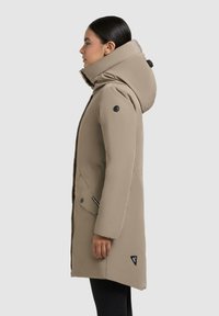 Beige geïsoleerde lange jas met hoge kraag, grote capuchon, zijzakken en zwarte hardwaredetails. Zachte textuur met een getailleerde snit.