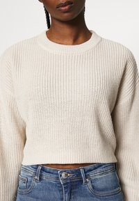 Lys beige strikket sweater med ribbet tekstur, bred rund hals og cropped design. Sammen med blå denim jeans med klassisk syning.