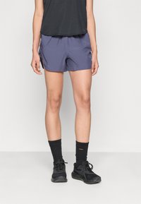 Pantalones cortos deportivos morados con textura acanalada, cinturilla elástica y aberturas en los laterales. Se llevan con una camiseta negra y calcetines y zapatos deportivos negros.