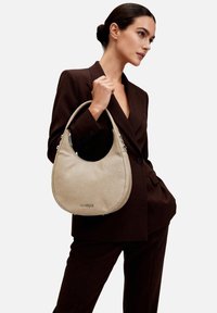 Donna in completo marrone scuro che tiene una borsa a mezzaluna beige con manico corto e logo argentato, in piedi davanti a uno sfondo bianco.