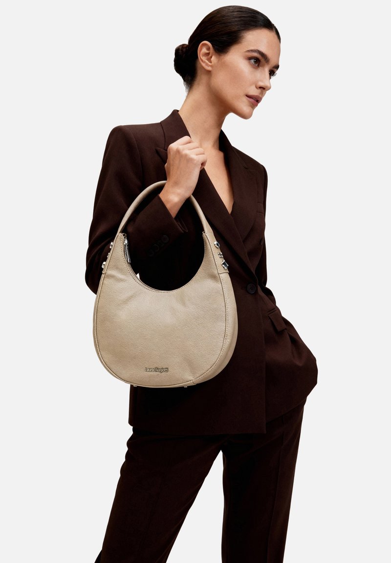 Donna in completo marrone scuro che tiene una borsa a mezzaluna beige con manico corto e logo argentato, in piedi davanti a uno sfondo bianco.