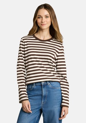 Donna con i capelli lunghi che indossa una maglietta a maniche lunghe a righe marroni e bianche e jeans blu, in piedi contro uno sfondo chiaro e uniforme.