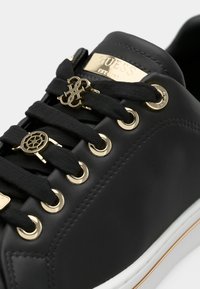 Baskets noires avec œillets dorés, plaque logo Guess dorée et breloques décoratives dorées sur lacets noirs, semelle blanche avec liseré doré.