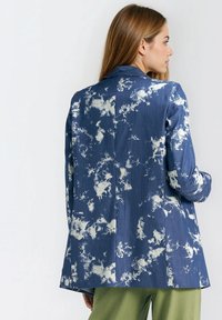 Granatowa marynarka w deseń przypominający białe chmury; o strukturze z gładką fakturą i pojedynczym rozporkiem z tyłu; w zestawieniu z zielonymi spodniami.