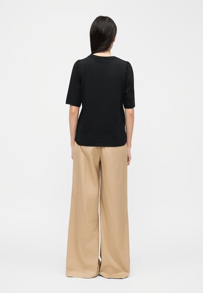 Femme aux longs cheveux noirs portant un haut noir à manches courtes et un pantalon beige à jambes larges, debout de dos sur un fond uni.