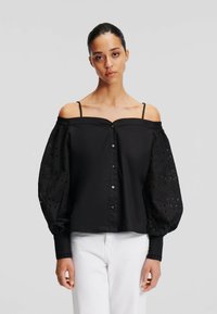 Donna che indossa una blusa nera a spalle scoperte con maniche a palloncino in pizzo e pantaloni bianchi, in piedi davanti a uno sfondo grigio chiaro uniforme.