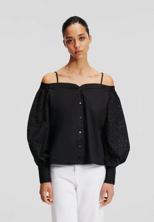 Vrouw in een zwart off-shoulder blouse met kanten ballonmouwen en witte broek, staand voor een effen lichtgrijze achtergrond.