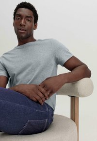 Lichtgrijze katoenen t-shirt met een ronde hals, gecombineerd met donkerblauwe jeans, zittend op een crème kleurige gestoffeerde stoel met houten armleuningen.