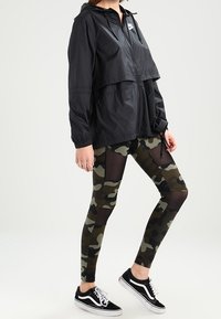 Musta vetoketjullinen hupullinen takki kerroksittain, yhdistettynä maastokuvioisiin leggingsseihin, joissa on verkkopaneeleita. Malli käyttää mustia lenkkareita.