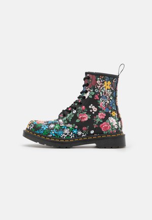 Sort snørestøvle med lyst, flerfarvet blomsterprint og gul syning på sålen, set fra siden mod en hvid baggrund.