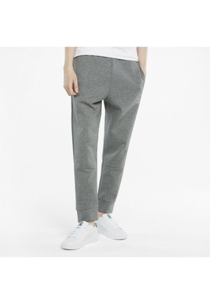 Personne portant un pantalon de survêtement gris et des baskets blanches, debout avec une main dans la poche sur un fond blanc uni.