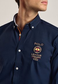 Camisa de botonadura azul marino hecha de algodón. Cuenta con botones blancos, logo bordado y un borde multicolor en el cuello. Diseño sencillo.