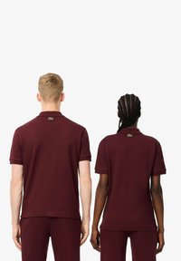 Bordowe koszulki polo wykonane z bawełny, z krótkimi rękawami, kołnierzykiem i logo z tyłu, noszone przez dwóch modeli od tyłu.