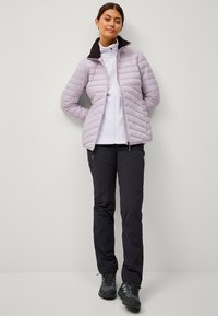 Giacca trapuntata viola chiaro con colletto nero, indossata sopra un maglione di pile bianco e pantaloni neri. L'outfit presenta tasche funzionali e stivali resistenti.