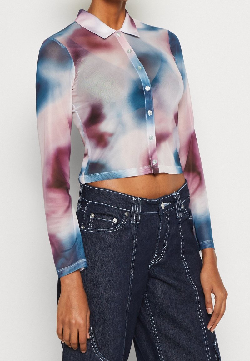 Femme portant un crop top à manches longues et à boutons avec un motif tie-dye rose, bleu et blanc, assorti à un jean taille haute bleu foncé.
