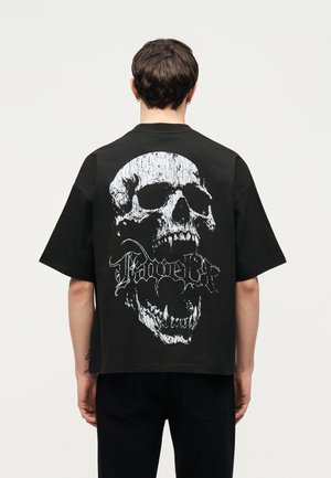 Person with short dark hair wearing a black shirt featuring a large white distressed skull graphic and stylized text on the back.