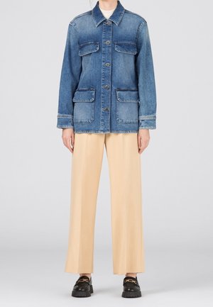 Persona que lleva una chaqueta de mezclilla azul abotonada con bolsillos delanteros, pantalones beige de pierna ancha y mocasines negros con detalle de cadena dorada.