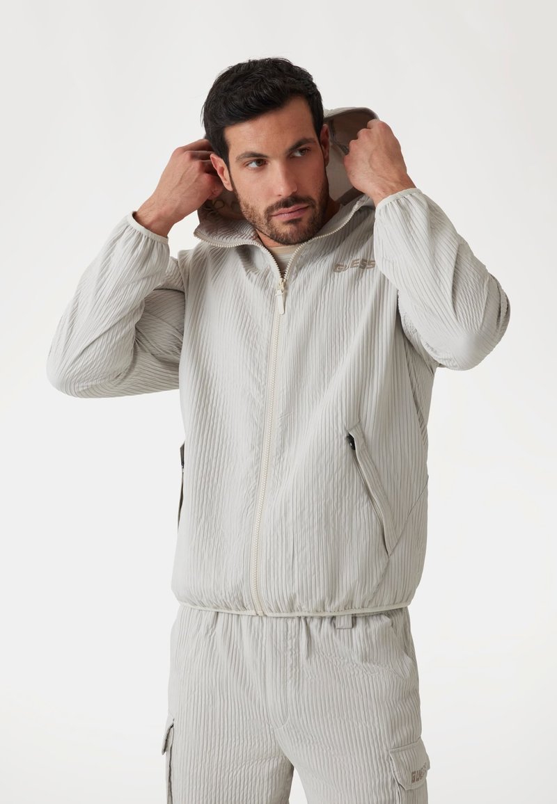 Giacca zip-up grigio chiaro con texture a coste verticali, cappuccio attaccato, tasche laterali e dettaglio del logo sul petto. Finitura del tessuto liscia.