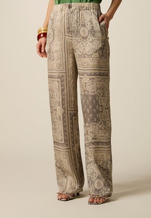 Donna che indossa pantaloni beige e marroni ampi con fantasia e vita elastica, abbinati a sandali con tacco a stampa serpente e braccialetti colorati.