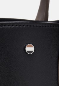 Szczegół czarnej skórzanej torebki z okrągłym metalowym akcentem ozdobionym poziomymi paskami w kolorach brązowym, białym i czarnym, widoczne zszywane krawędzie.